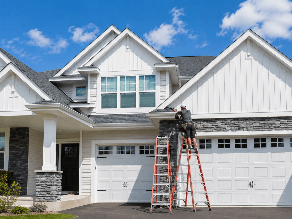Worker on ladders performing gutter maintenance on a large modern suburban home.