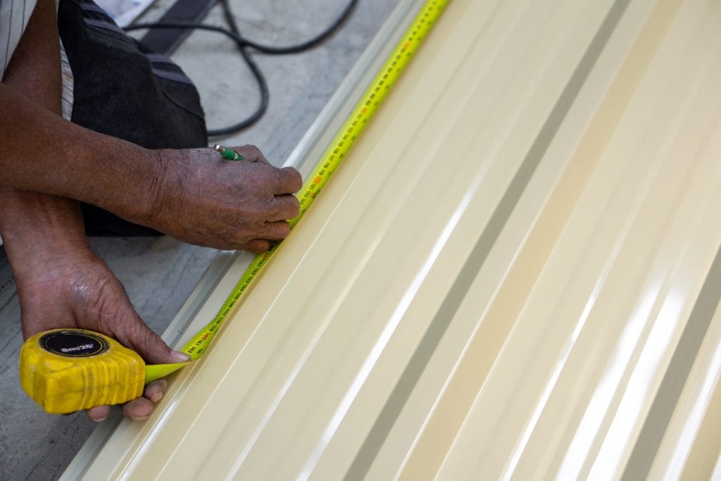metal roof installation okc A person is using a yellow measuring tape to measure sheet metal in a workshop. The focus is on precise measurement for future task.